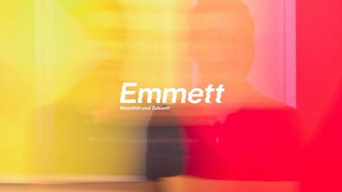 Schriftgrafik mit dem Text „Emmett. Mobilität und Zukunft.“ Im Hintergrund sind ein gelb-roter Farbverlauf sowie die Spiegelung einer Person zu erkennen.