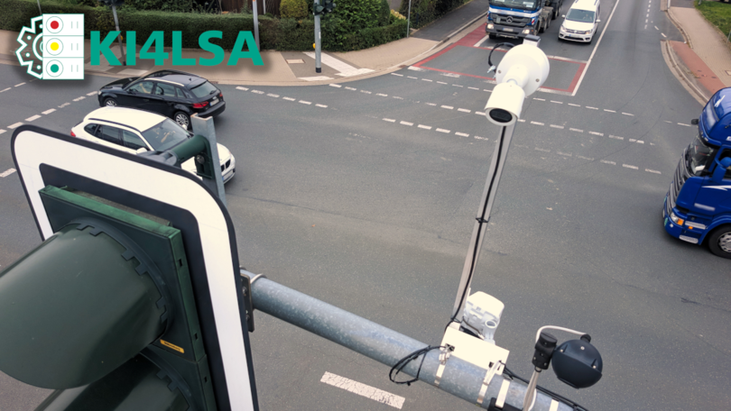 Straßenkreuzung mit verschiedenen Fahrzeugen. Im Vordergrund sind Sensoren und Kameras an einer Lichtsignalanlage angebracht.