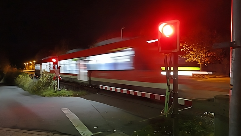Ein Bahnübergang mit geschlossener Schranke und roter Lichtsignalanlage wird von einer Regionalbahn durchfahren.