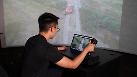 Anwendungsbeispiel für das Projekt SKADRO: Eine Person blickt auf einen Laptop-Bildschirm und steuert eine Drohne. Im Hintergrund wird das Kamera-Bild der Drohne an die Wand projiziert. Es zeigt die Luftaufnahme eines Fahrzeuges auf einer Straße.