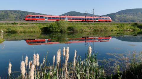 Eine Regionalbahn fährt durch eine Landschaft, im Vordergrund spiegelt sich die Bahn in einem Gewässer