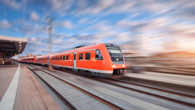 Eine Regionalbahn durchfährt einen Bahnhof. Am linken Bildrand ist ein leerer Bahnsteig zu sehen. Der Fokus liegt auf dem Zug selbst. Der Rest des Bildes ist unscharf und deutet dadurch die Geschwindigkeit der Regionalbahn an.