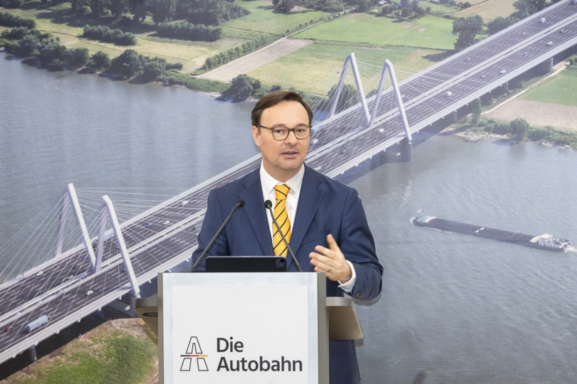 Oliver Luksic spricht am Rednerpult. Im Hintergrund ist eine Visualisierung der zukünftigen, fertiggestellten Rheinbrücke zu sehen.