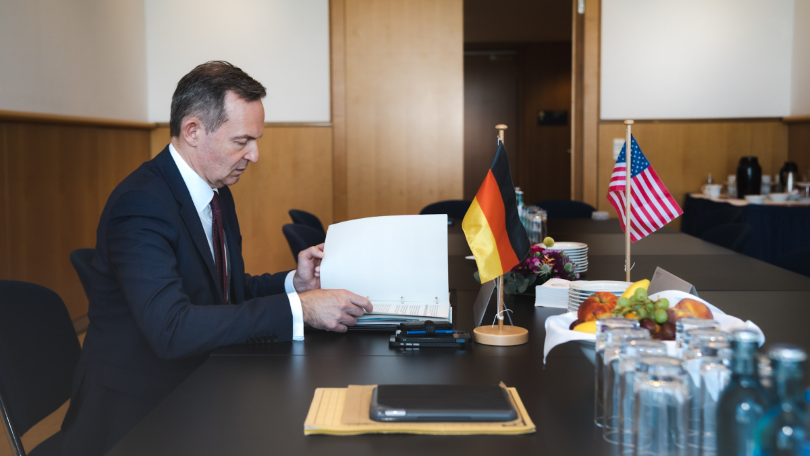 Bundesminister Dr. Volker Wissing beim Weltverkehrsforum 2022. Der Minister sitzt an einem Konferenztisch und liest Dokumente. Auf dem Tisch stehen die deutsche und die US-amerikanische Flagge sowie eine Obstschale und mehrere Gläser.