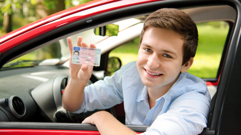 Junger Mann der seinen Führerschein aus einem Auto heraus lächelnd zeigt